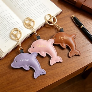 May include: Three dolphin-shaped keychains in lavender, light pink, and brown. Each keychain features a gold-tone metal ring and clasp attached by a black strap. The names Jesse, Maria, and Dylan are embossed in gold on the respective dolphin figures.