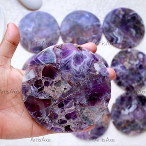 May include: A collection of round amethyst coasters. The coasters are a mix of deep purple, white, and gray. The ArtisAxe logo is visible on the coasters. The coasters are made of natural stone.
