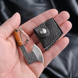 May include: A miniature axe keyring featuring a wooden handle, a patterned metal blade, and a silver-toned ball chain. Accompanying the axe is a small, black, textured leather-like pouch with a silver snap button closure for storage and protection.