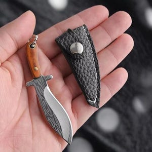 Può includere: Un coltello in miniatura con manico in legno marrone chiaro, lama argentata con finitura a motivi scuri e un piccolo anello metallico sulla parte superiore. È accompagnato da un fodero nero testurizzato con chiusura a bottone argentato.