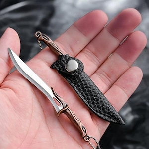 Può includere: Un ciondolo a forma di pugnale in miniatura con un manico in metallo color rame dai dettagli complessi e una lama color argento. Il pugnale è accompagnato da un fodero in similpelle nera testurizzata, fissato con un piccolo bottone a pressione argentato.