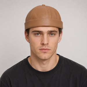 Puede incluir: Un gorro tipo docker de color tostado confeccionado en un material liso similar al cuero. El gorro presenta una corona redondeada de varios paneles con costuras verticales visibles y un ala ancha doblada que rodea la cabeza por completo.