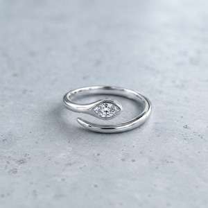 May include: A silver-coloured bypass ring featuring a single marquise-cut clear gemstone set at one end of the open band. The metal has a smooth, polished finish, and the design curves around to create a sleek, continuous shape for the finger.