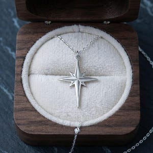 May include: A silver-toned necklace featuring a four-pointed star pendant with elongated rays. The pendant is centred within a dark wooden box lined with cream-coloured velvet, showcasing a delicate chain that extends from the star charm.