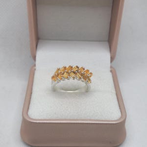 Puede incluir: Un anillo de plata con dos filas de piedras preciosas naranjas de corte baguette dispuestas en un patrón escalonado y superpuesto. El anillo se muestra dentro de un joyero de color rosa claro con bisagras y un forro interior de color blanco.