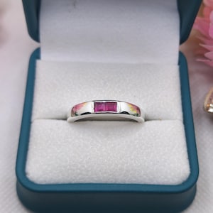 Puede incluir: Un anillo de tono plateado con dos piedras preciosas de color rosa vibrante y corte cuadrado, engastadas una al lado de la otra en el centro de la banda. El anillo se presenta en un joyero azul oscuro con un forro interior blanco texturizado.
