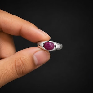 May include: A silver ring featuring a smooth, oval-shaped, deep pinkish-red gemstone set in the center. The metal band has a polished, reflective finish and a simple, solid design that tapers slightly toward the back of the ring setting.