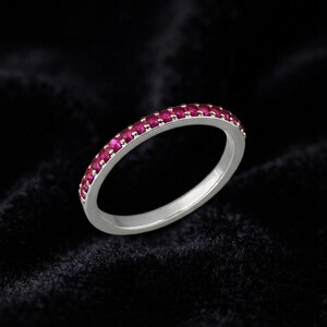 Puede incluir: Un anillo de banda en tono plateado que presenta una fila continua de pequeñas piedras preciosas redondas de color rojo rosado vibrante, engastadas a lo largo de la mitad superior de la banda metálica con un acabado pulido y liso.