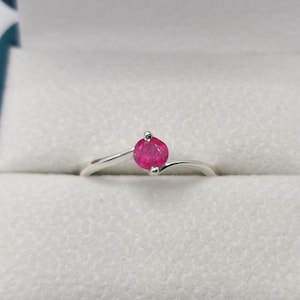 Op de afbeelding: Een zilveren ring met een enkele, rond geslepen, felroze edelsteen in een ringband in bypass-stijl. De metalen band buigt zich soepel rond de centrale steen, die op zijn plaats wordt gehouden door een kleine zetting met pootjes.