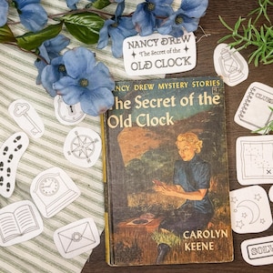 May include: A vintage copy of "The Secret of the Old Clock" by Carolyn Keene, with a woman on the cover, surrounded by detective-themed stickers. The stickers include a key, compass, and magnifying glass. Blue flowers and greenery are also present.