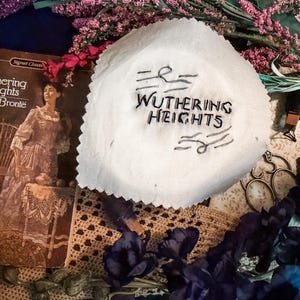 Puede incluir: Una portada de libro vintage de "Cumbres Borrascosas" de Emily Brontë, con una mujer con un vestido. Un tejido blanco de forma irregular con el título bordado en hilo negro está en primer plano. También se ven flores moradas y encaje.