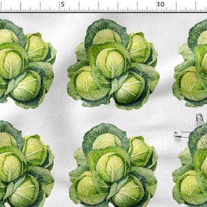 May include: A white fabric featuring a repeating pattern of green cabbage heads. A metal safety pin is attached to the fabric near the bottom right. A ruler along the top and left edges shows the scale in centimetres, with the pattern repeating.