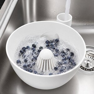 Könnte beinhalten: Eine weiße Kunststoff-Siebschüssel mit einem mittig erhöhten Abflusskegel, gefüllt mit frischen Blaubeeren, die in Wasser eingetaucht sind. Die Schüssel hat ein glattes, abgerundetes Design und lässt Wasser durch den zentralen Abfluss ab.