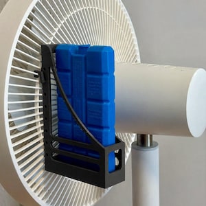 Könnte beinhalten: Ein weißer Standventilator mit einer schwarzen Kunststoffhalterung an der Vorderseite. Ein rechteckiges blaues Kühlakku ist in der Halterung befestigt und direkt vor den Ventilatorflügeln positioniert, um den Luftstrom beim Durchgang zu kühlen.
