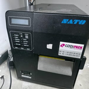 May include: A black SATO industrial label printer featuring a digital display, a control panel with buttons labeled Power, Line, Feed, Print, Offset, Pitch, and Display, and a white sticker with the text CODiPACK and contact phone numbers.