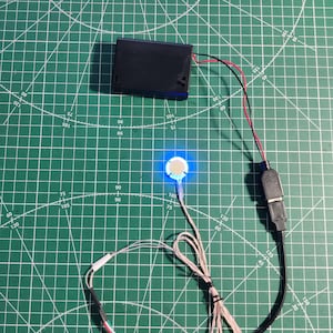 May include: A black rectangular battery pack connected by thin red and black wires to a small circular component emitting a bright blue light. This component is linked to a black USB connector and a bundle of thin white wires with white tape.