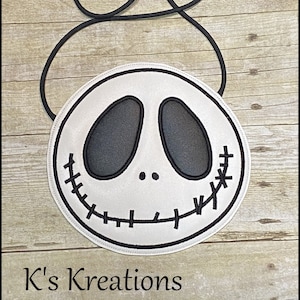 Pu&ograve; includere: Una borsa a tracolla rotonda bianca con un design a volto di Jack Skellington nero. La borsa ha dettagli cuciti neri per la bocca e gli occhi e una tracolla in corda nera. Il testo "K's Kreations" &egrave; in basso.