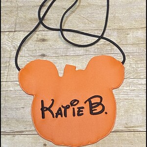 May include: Orange crossbody bag shaped like a mouse head, with a black cord strap. The name "Katie B." is written in black script on the front. The words "K's Kreations" are at the bottom.