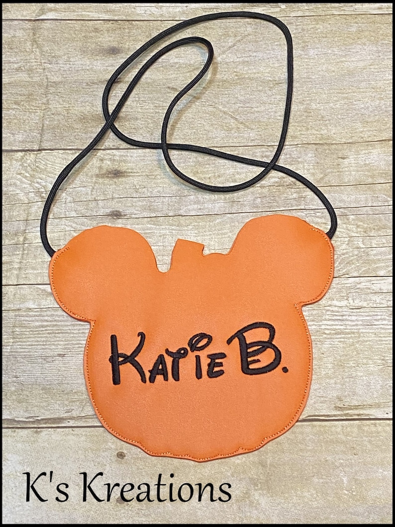 May include: Orange crossbody bag shaped like a mouse head, with a black cord strap. The name "Katie B." is written in black script on the front. The words "K's Kreations" are at the bottom.