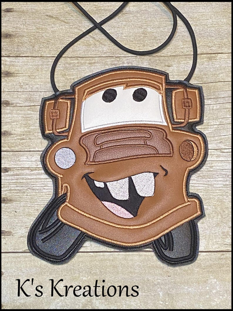 May include: A brown and tan crossbody bag shaped like the character Mater from the movie Cars. The bag has a black strap and features Mater's face with large teeth and a smiling mouth. The text "K's Kreations" is at the bottom.