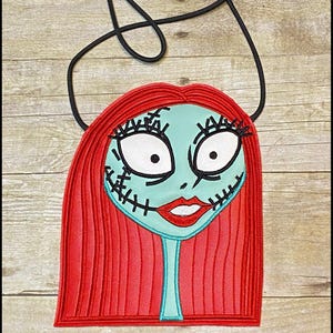 Pu&ograve; includere: Una borsa a tracolla con un volto stilizzato, con capelli rossi, un viso turchese e dettagli cuciti. La borsa ha una tracolla nera e il testo "K's Kreations" in basso. Il design ricorda un personaggio di un film d'animazione.