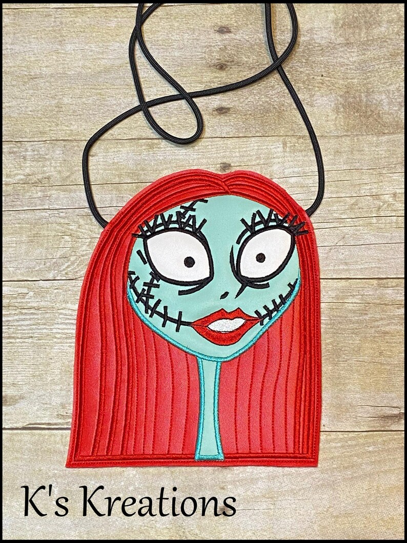Pu&ograve; includere: Una borsa a tracolla con un volto stilizzato, con capelli rossi, un viso turchese e dettagli cuciti. La borsa ha una tracolla nera e il testo "K's Kreations" in basso. Il design ricorda un personaggio di un film d'animazione.