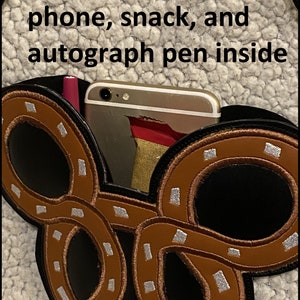 May include: Black purse with a pretzel design, containing a phone, snack, and pen. The purse has a black strap and a brown and silver pretzel design. Text on the image reads: "Purse shown with: phone, snack, and autograph pen inside."