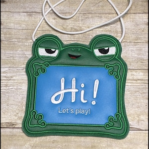May include: A green frog-shaped lanyard with a blue center, featuring the text "Hi! Let's play!" in white script. The frog has sleepy eyes and a red smiling mouth. A white cord is attached for wearing.