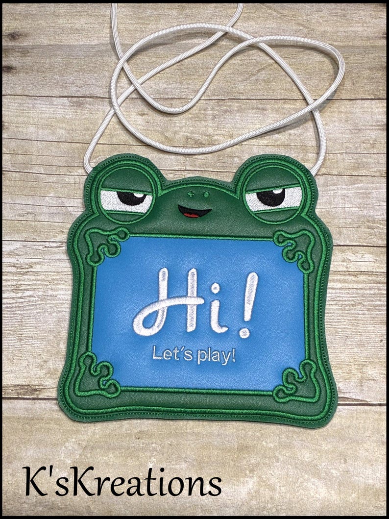 May include: A green frog-shaped lanyard with a blue center, featuring the text "Hi! Let's play!" in white script. The frog has sleepy eyes and a red smiling mouth. A white cord is attached for wearing.