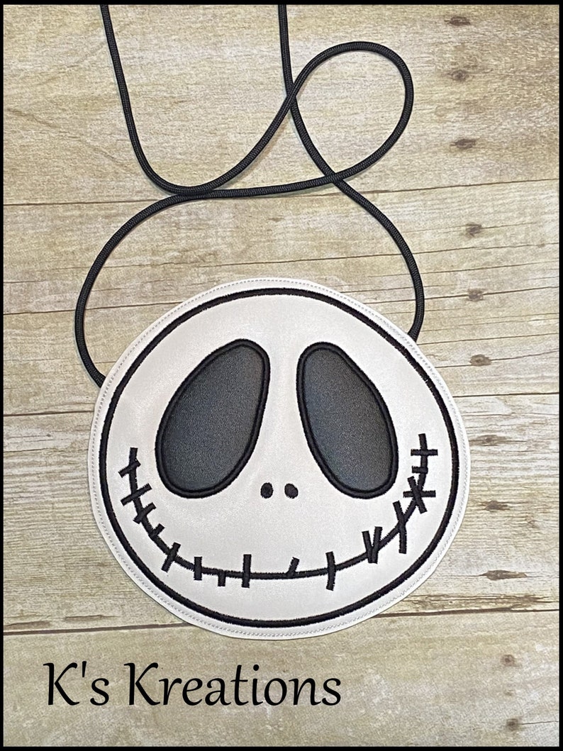 Pu&ograve; includere: Una borsa a tracolla rotonda bianca con un design a volto di Jack Skellington nero. La borsa ha dettagli cuciti neri per la bocca e gli occhi e una tracolla in corda nera. Il testo "K's Kreations" &egrave; in basso.