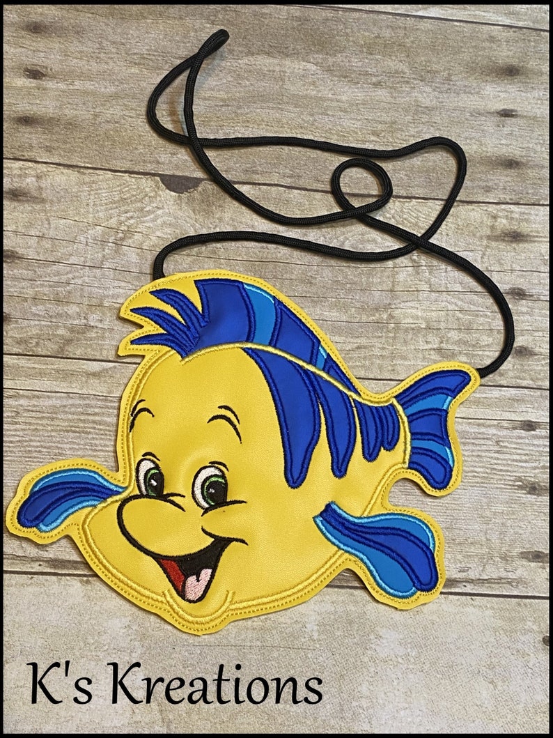 May include: A yellow and blue Flounder-shaped crossbody bag with a black strap. The bag features a smiling Flounder design with green eyes and a red mouth. The text "K's Kreations" is at the bottom of the image.
