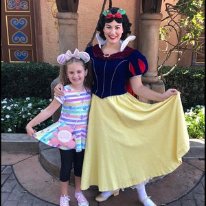 May include: A young girl and a woman dressed as Snow White pose for a photo. The girl wears a colorful striped dress, black leggings, and pink sneakers. Snow White wears a blue and yellow dress with a red cape. The text "K's Kreations" is at the bottom.