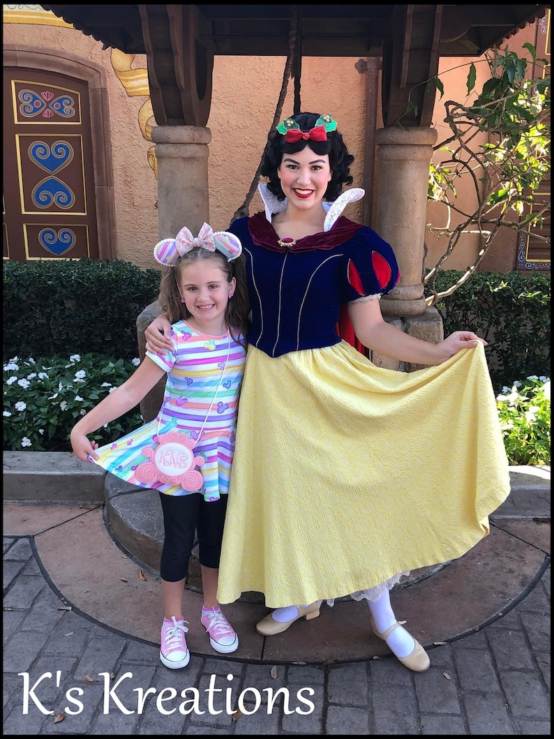 May include: A young girl and a woman dressed as Snow White pose for a photo. The girl wears a colorful striped dress, black leggings, and pink sneakers. Snow White wears a blue and yellow dress with a red cape. The text "K's Kreations" is at the bottom.