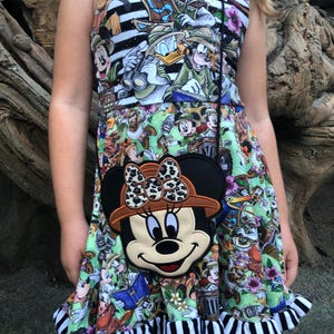 May include: A sleeveless dress with a colorful Disney character print, featuring a black and white striped bodice and a ruffled hem. A Minnie Mouse-shaped crossbody bag with a safari hat and leopard print bow is worn over the dress.