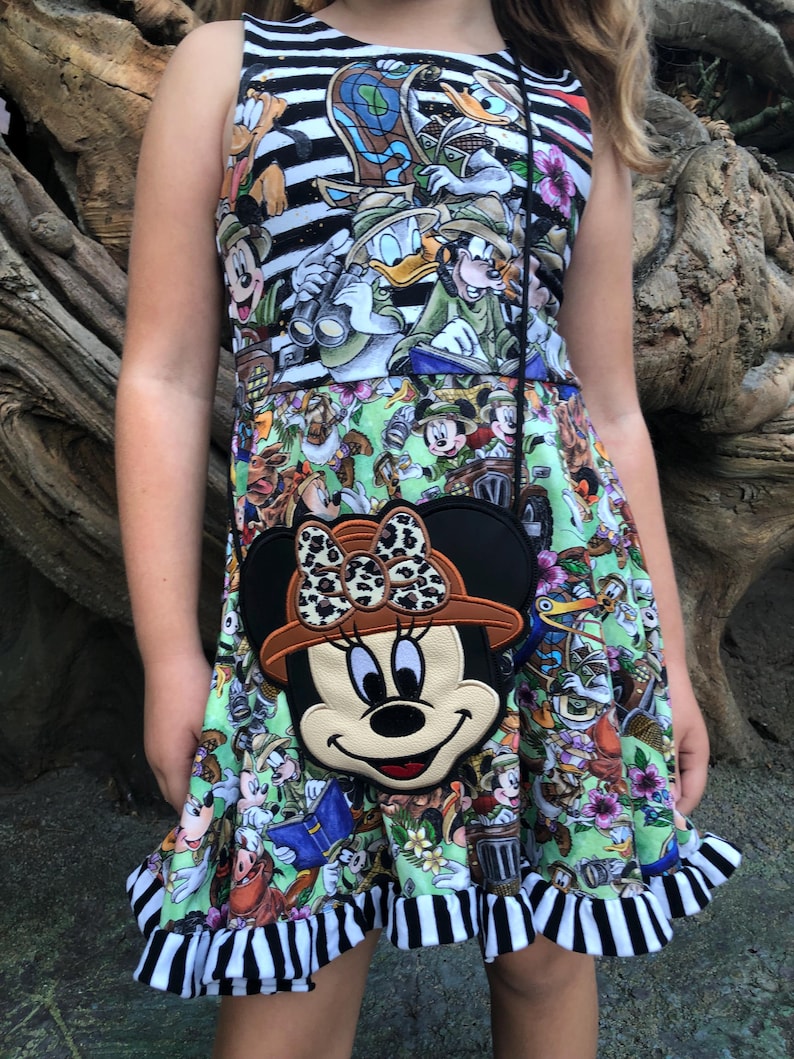 May include: A sleeveless dress with a colorful Disney character print, featuring a black and white striped bodice and a ruffled hem. A Minnie Mouse-shaped crossbody bag with a safari hat and leopard print bow is worn over the dress.
