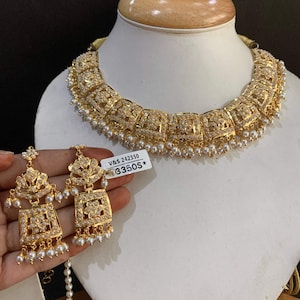 Puede incluir: Un conjunto de collar y pendientes a juego en tono dorado. El collar presenta eslabones cuadrados con detalles intrincados, adornados con pequeñas piedras transparentes y perlas colgantes. Los pendientes reflejan el diseño del collar. El conjunto se exhibe en un maniquí.