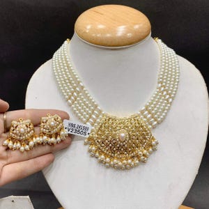 Pode incluir: Um colar de pérolas brancas de várias camadas com um grande pingente central dourado adornado com padrões intrincados de filigrana e pequenas pérolas pendentes. Brincos dourados a condizer com detalhes semelhantes de filigrana e pérolas.