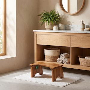 May include: A low, rectangular wooden step stool with a curved top surface and arched side supports, positioned on a white rectangular bath mat in front of a light oak bathroom vanity cabinet with open shelving and woven storage baskets.