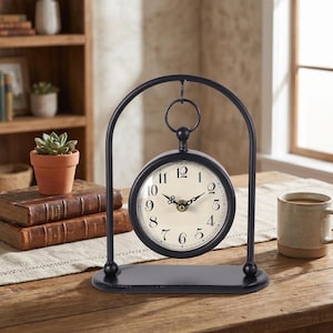 May include: A black metal desk clock with a cream-colored face and black numerals. The clock is supported by a curved metal frame and a rectangular base. The clock's hands are black and the clock is sitting on a wooden table.