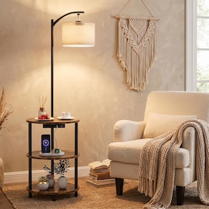 May include: A black metal floor lamp with a beige fabric drum shade and a three-tier circular side table. The table features a dark wood-grain finish, a built-in power strip with USB ports, and three shelves holding small potted plants and a phone.