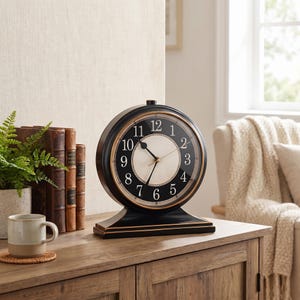 May include: A black and gold-trimmed mantel clock with a white face and black numerals. The clock has a round shape and sits on a rectangular base. The clock is approximately 10 inches tall.