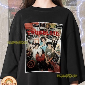 Puede incluir: Una camiseta negra de cuello redondo con un gran estampado gráfico rectangular en el pecho. El gráfico muestra un collage de varios carteles de conciertos con el texto rojo TRUEBLOOD superpuesto. Cuatro personas aparecen en el centro del collage.