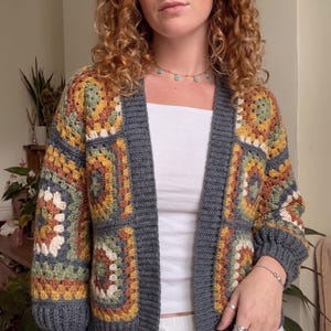 Può includere: Un cardigan a maniche lunghe realizzato con toppe all'uncinetto granny square multicolori nei toni del giallo senape, verde salvia, crema e arancione bruciato, rifinito con bordi, polsini e apertura frontale a coste grigio scuro.
