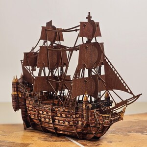 May include: A detailed wooden model of a multi-masted sailing ship featuring dark brown laser-cut panels, intricate rigging, multiple square sails, and a decorative stern structure. The ship is constructed from flat interlocking pieces.