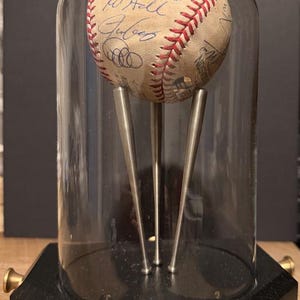 May include: A baseball with signatures, encased in a glass dome. The baseball is displayed on a black wooden base with brass-colored accents. The baseball has red stitching and is supported by three silver metal rods.