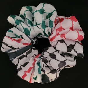 Puede incluir: Un coletero de tela circular con un patrón geométrico repetitivo en blanco con secciones en verde, rojo y negro. El material tiene una apariencia tejida y texturizada con pliegues fruncidos que crean una forma circular voluminosa y ondulada.