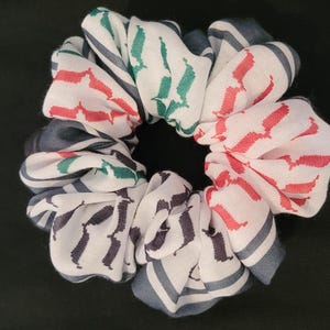 May include: A fabric hair scrunchie featuring a white base with a repeating pattern of red, dark green, and dark grey geometric shapes and lines. The accessory has a gathered, ruffled texture throughout, creating a circular shape with a centre opening.