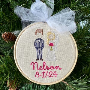 May include: A white and brown embroidered ornament with a couple, a groom in a blue suit and a bride in a white dress, with the text "Nelson 8-17-24" in pink thread.