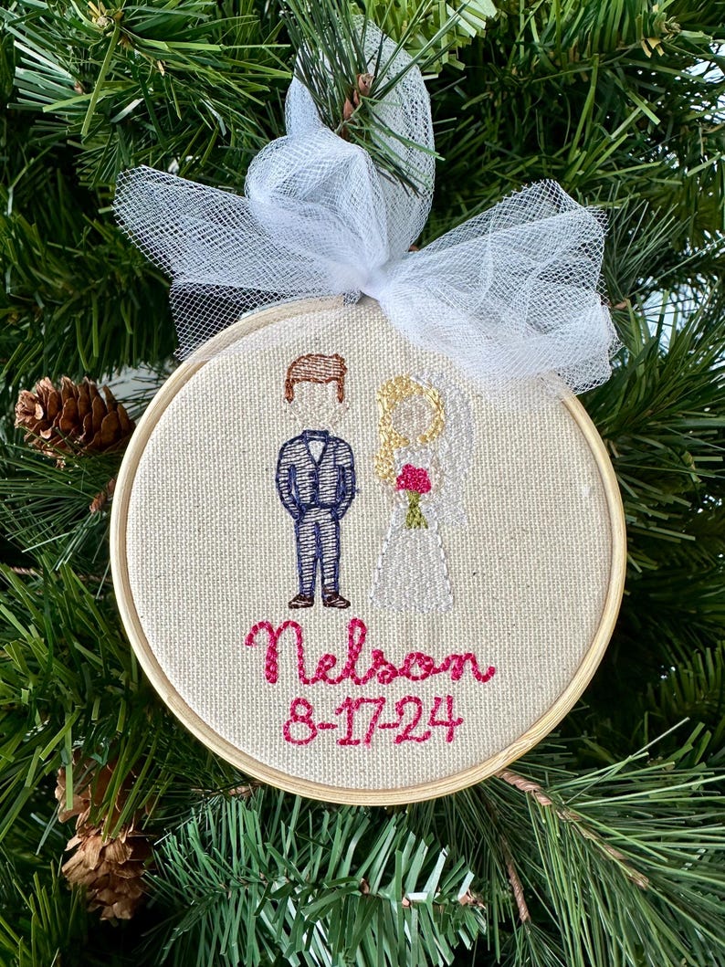 May include: A white and brown embroidered ornament with a couple, a groom in a blue suit and a bride in a white dress, with the text "Nelson 8-17-24" in pink thread.