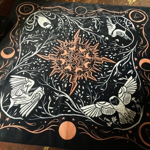 May include: Black fabric tapestry with a white and peach design. The design features a sun with rays, two birds, and a crescent moon. The tapestry has a border of peach and white.